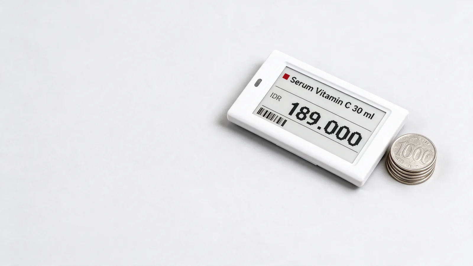 A white electronic shelf label lying flat beside a small stack of Indonesian rupiah coins.